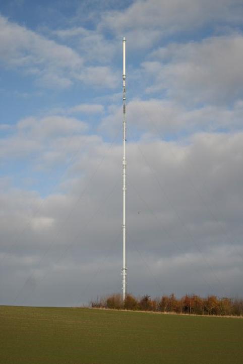 A colour photo of a white TV mast against a cloudy sky. The mast is very tall, with lots of cables tensioned between it and the ground. The mast is in a field, with some trees at the base.